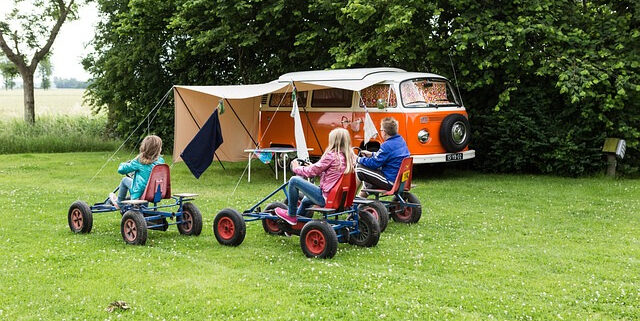 Pressearbeit in der Ferienzeit. Campingbus mit Zelt. Davor Kinder auf Gokarts auf Wiese.