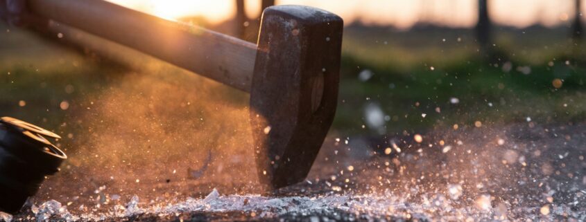 Ein schwerer Hammer trifft im sonnigen Gegenlicht auf einen Amboss und zerschmetter Glas, dass es funkelt.