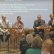 Auf einer Bühne sitzen 5 Personen auf Barhockern vor einem Publikum. Podiumsdiskussion.