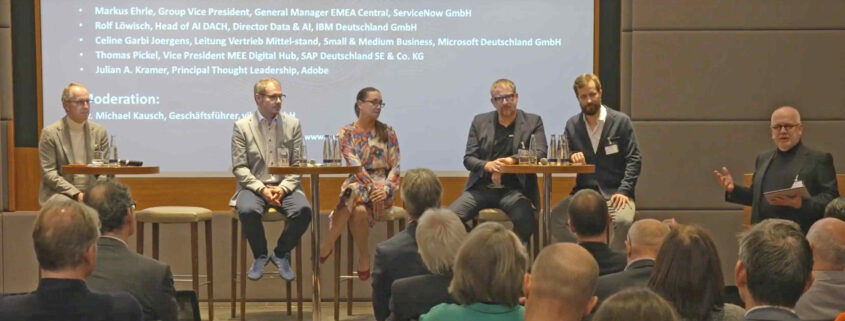 Auf einer Bühne sitzen 5 Personen auf Barhockern vor einem Publikum. Podiumsdiskussion.