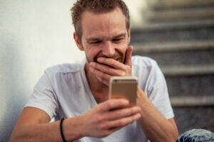 Mann hat ein Smartphone in der Hand. Er hat die Hand vor dem Mund und lacht.